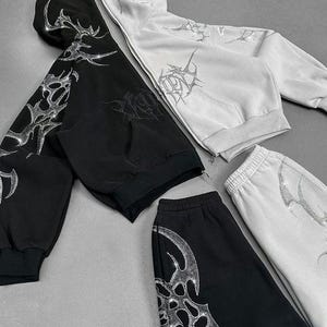 Op de afbeelding: Zwart en wit bijpassend trainingspak. De set bevat een hoodie met rits en een joggingbroek, beide versierd met een zilverkleurig, op strass lijkend tribaal ontwerp. De hoodie heeft een capuchon en voorzakken. De joggingbroek heeft een elastische tailleband.