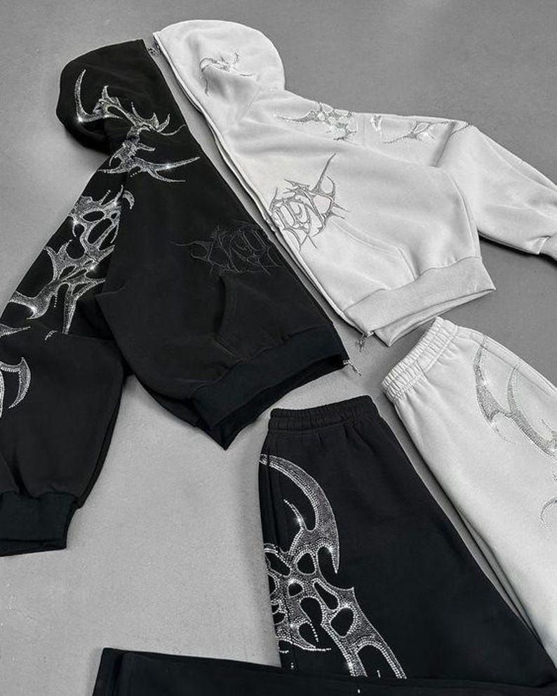 Op de afbeelding: Zwart en wit bijpassend trainingspak. De set bevat een hoodie met rits en een joggingbroek, beide versierd met een zilverkleurig, op strass lijkend tribaal ontwerp. De hoodie heeft een capuchon en voorzakken. De joggingbroek heeft een elastische tailleband.
