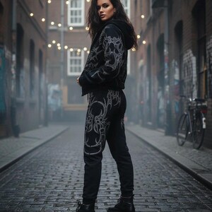 Op de afbeelding: Zwart jack met capuchon en bijpassende broek met een zilveren tribal design. De outfit bevat zwarte combat boots. De persoon staat op een geplaveide straat.