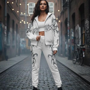 Op de afbeelding: Witte trainingspak met een donkergrijs tribaal ontwerp, bestaande uit een capuchonjas met rits en bijpassende joggingbroek. De persoon draagt een witte crop top en zwarte laarzen. De achtergrond is een steegje met bakstenen en lichtslingers.