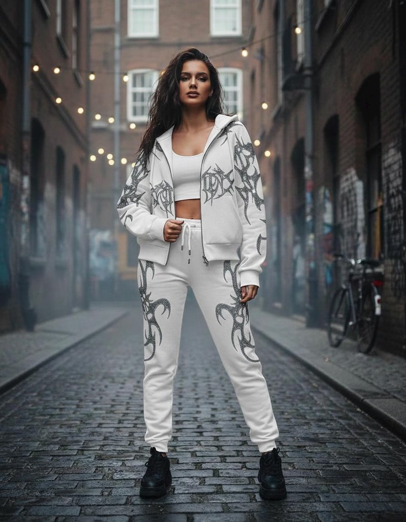 Op de afbeelding: Witte trainingspak met een donkergrijs tribaal ontwerp, bestaande uit een capuchonjas met rits en bijpassende joggingbroek. De persoon draagt een witte crop top en zwarte laarzen. De achtergrond is een steegje met bakstenen en lichtslingers.