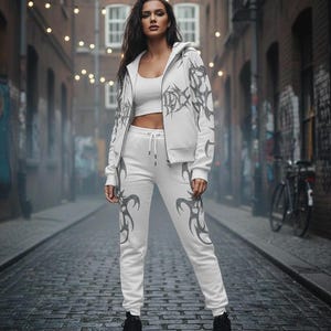 Op de afbeelding: Witte outfit bestaande uit een hoodie, crop top en joggingbroek. De kleding heeft een donkergrijs tribaal ontwerp. Het model draagt zwarte laarzen. De achtergrond is een geplaveide straat met bakstenen gebouwen en lichtslingers.