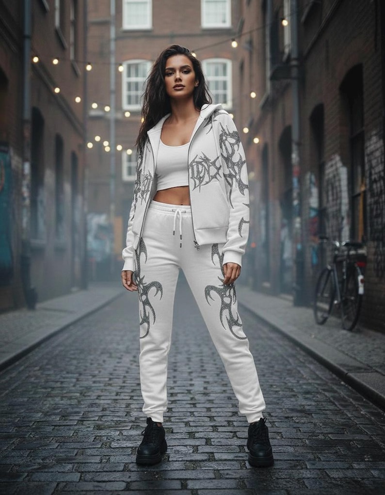 Op de afbeelding: Witte outfit bestaande uit een hoodie, crop top en joggingbroek. De kleding heeft een donkergrijs tribaal ontwerp. Het model draagt zwarte laarzen. De achtergrond is een geplaveide straat met bakstenen gebouwen en lichtslingers.
