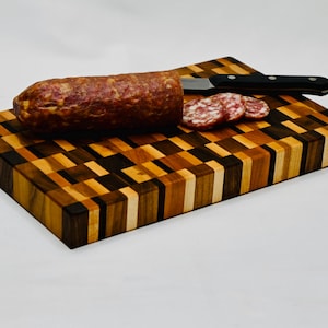 Puede incluir: Una tabla de cortar de madera con un patrón de tablero de ajedrez de varios tonos de madera. Una salchicha entera y salami en rodajas están en la tabla, con un cuchillo con mango negro. La tabla es rectangular y está sobre una superficie blanca.