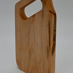 Op de afbeelding: Een lichtgekleurde houten snijplank met een handvat. De plank is rechthoekig met afgeronde hoeken en een gladde afwerking. Het handvat heeft een grote, ovale opening. De houtnerf is zichtbaar en de plank lijkt uit één stuk hout te bestaan.