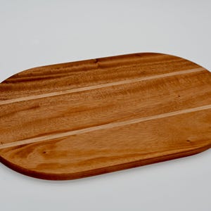 May include: Oval-shaped wooden serving board with a warm brown tone. The board has a smooth surface with visible wood grain and two parallel grooves. This decorative board is ideal for serving appetizers or charcuterie.