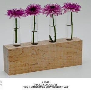 Op de afbeelding: Een houten bloemenvaas met vier heldere glazen buizen, elk met een paarse chrysant. De vaas is gemaakt van curly maple en afgewerkt met watergedragen satijn polyurethaan. Het Grain Craft Lab-logo staat linksonder.