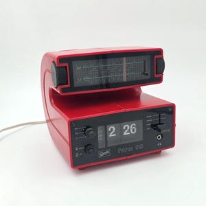 Può includere: Una radiosveglia in plastica rossa con display digitale a palette che mostra le 2:26. Il dispositivo presenta un sintonizzatore radio analogico montato superiormente con una manopola nera, un pannello frontale con manopole e la scritta Quartz form 99.