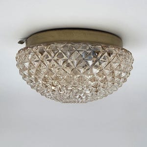 May include: A flush mount ceiling light fitting featuring a dome-shaped, clear glass shade with a textured, diamond-cut geometric pattern. The glass shade is secured by a circular, brushed brass-toned metal base plate at the top.