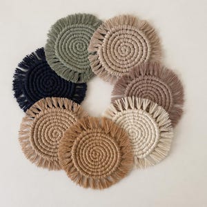 Könnte beinhalten: Eine Sammlung von acht handgefertigten Makramee-Untersetzern in verschiedenen neutralen Farben, darunter Beige, Braun und Marineblau. Jeder Untersetzer hat ein kreisförmiges Design mit einem Fransenrand, ideal für Getränke.