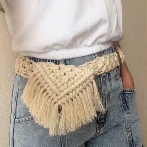 May include: A cream-colored macrame belt featuring a triangular front pouch design with long, dangling fringe details. The belt is constructed from thick, knotted cord and includes a metallic buckle closure, worn over light blue denim jeans.