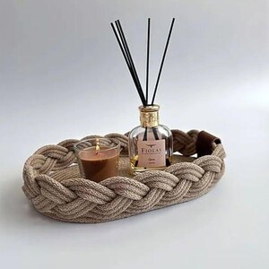 Könnte beinhalten: Dekorative ovale Ablage aus geflochtenem Seil in Beige. Sie enthält eine Glas-Kerze, eine Diffusorflasche mit Stäbchen und ein Etikett mit der Aufschrift „FIOLAS“. Ein stilvolles Wohnaccessoire.