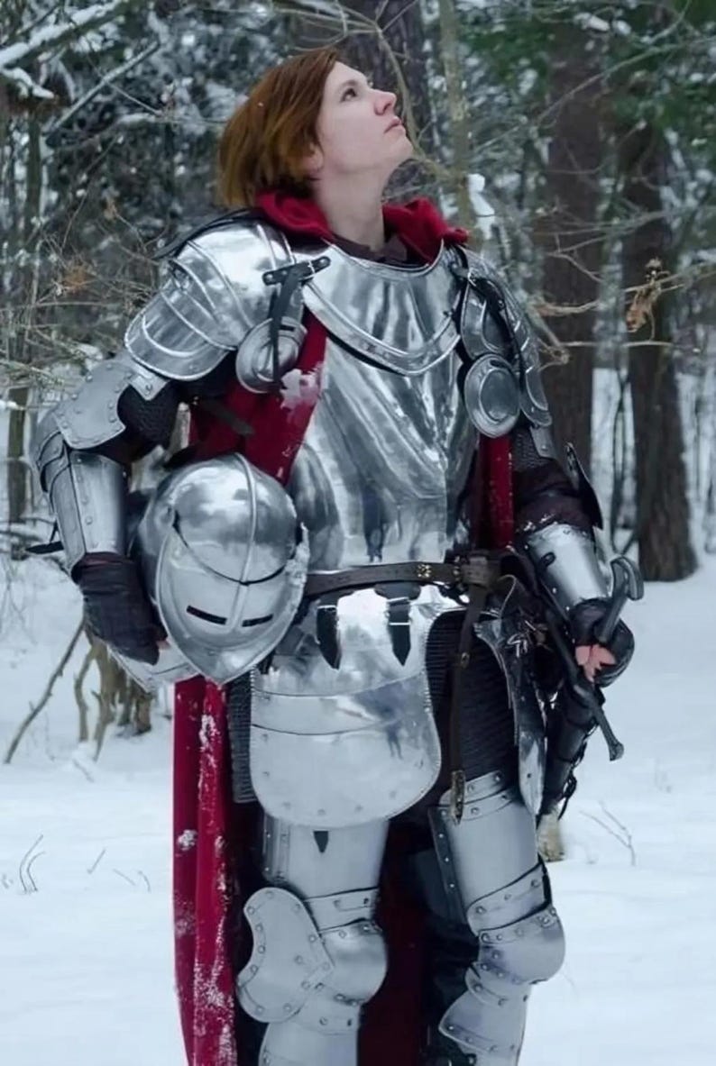May include: A person wearing full plate armor, including a helmet, chest plate, and leg protection. The armor is silver-colored and reflects the light. A red cape is visible under the armor. The person is standing in a snowy forest.