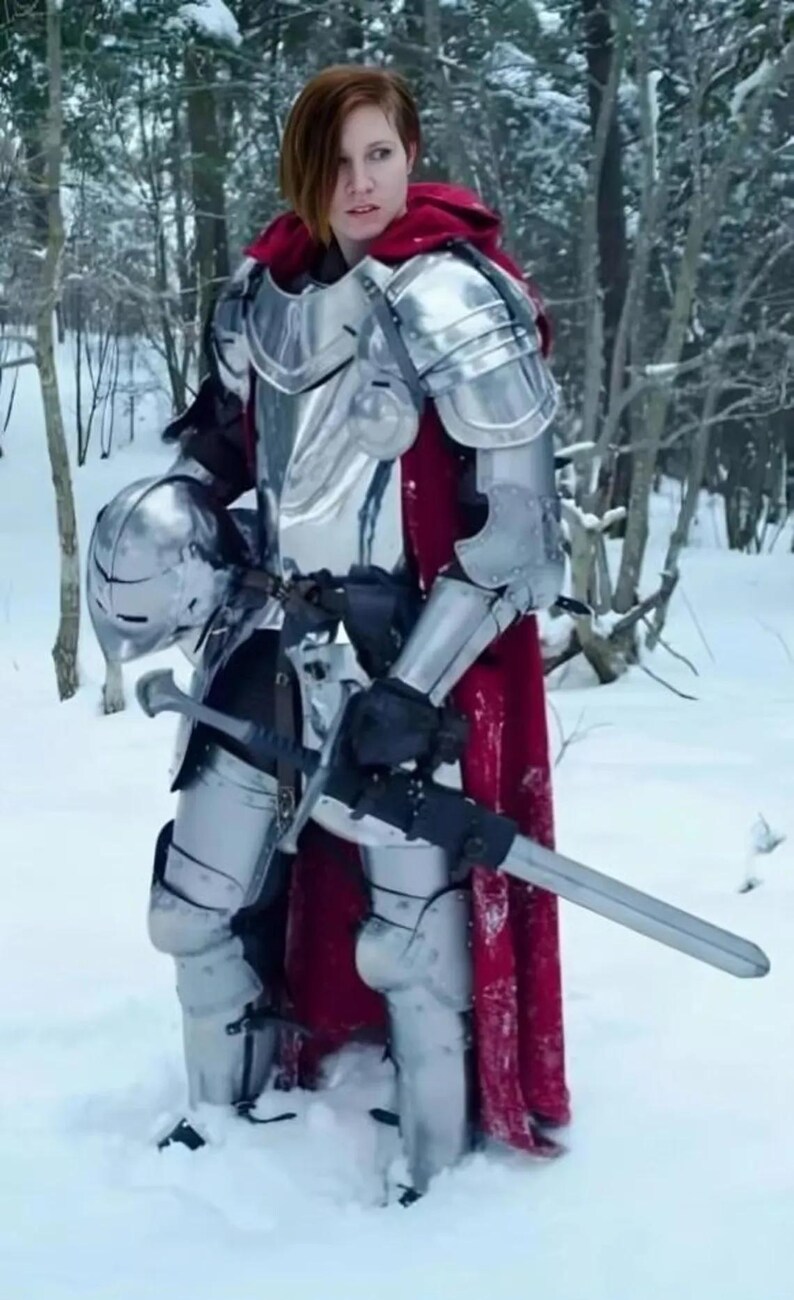 May include: A person wearing full plate armor, a red cape, and a helmet, holding a sword in a snowy forest. The armor is silver and reflective. The person is standing in deep snow.