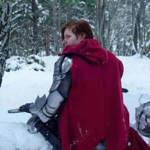 May include: A person wearing a red hooded cloak and silver armor, sitting in a snowy forest. The armor covers the shoulders and arms. A helmet rests in the snow. The scene is set in a winter landscape with snow-covered trees.