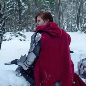 May include: A person wearing a red hooded cloak and silver armor, sitting in a snowy forest. The armor includes shoulder and arm guards. A helmet rests in the snow nearby. The scene is set in a winter landscape with snow-covered trees.