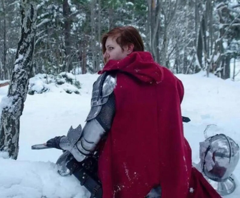 May include: A person wearing a red hooded cloak and silver armor, sitting in a snowy forest. The armor includes shoulder and arm guards. A helmet rests in the snow nearby. The scene is set in a winter landscape with snow-covered trees.