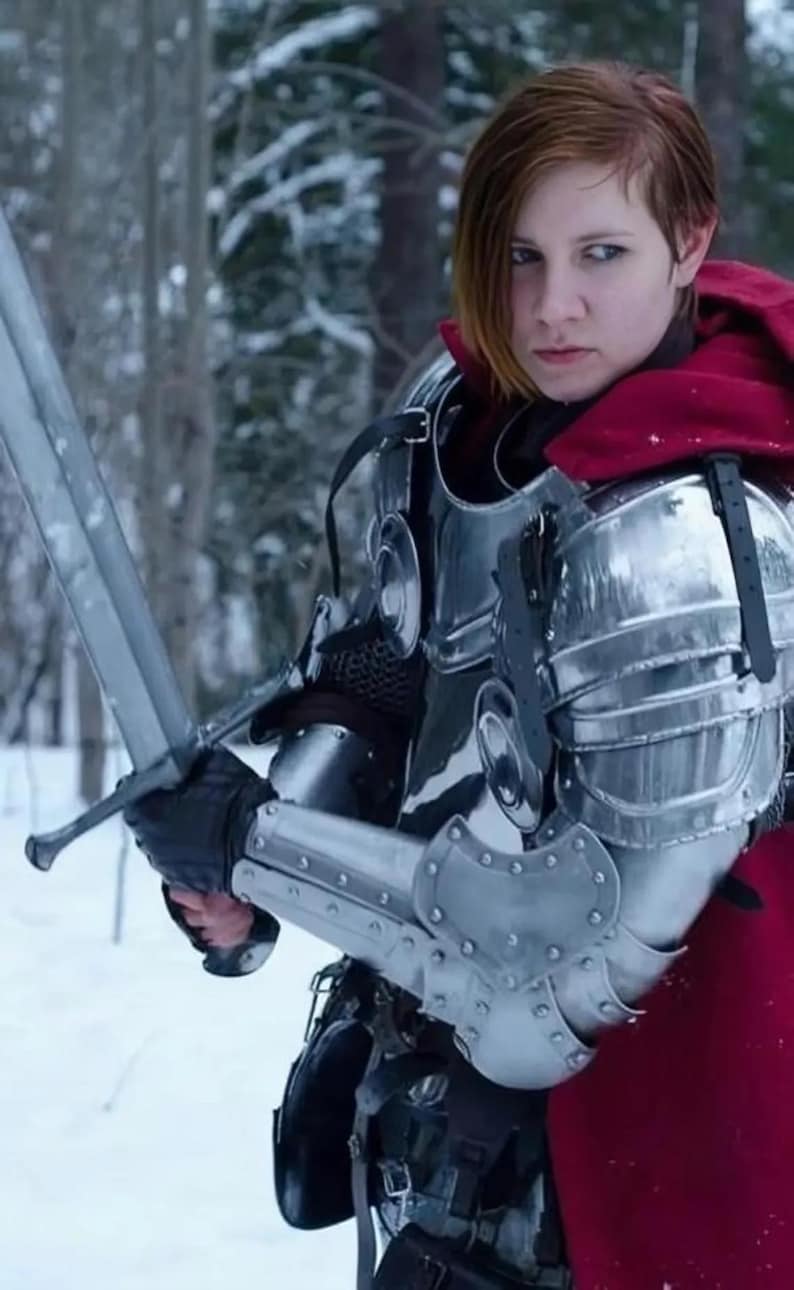 May include: A person in medieval armor, holding a long sword. The armor is silver-colored and reflects the light. A red cloak is draped over the shoulders. The background is a snowy forest.
