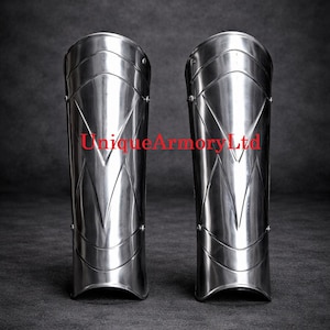 Pu&ograve; includere: Un paio di schinieri in acciaio lucido caratterizzati da un design a cresta verticale a forma di V e rivetti metallici sui lati. Il testo UniqueArmoryLtd &egrave; stampato in rosso al centro di entrambi i pezzi di armatura per la protezione delle gambe.