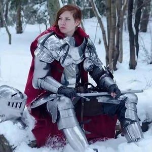 May include: A person wearing silver metal armor and a red cape sits in a snowy forest. The armor includes a helmet, chest plate, shoulder guards, arm and leg protection. A sword is held in the person's hands.