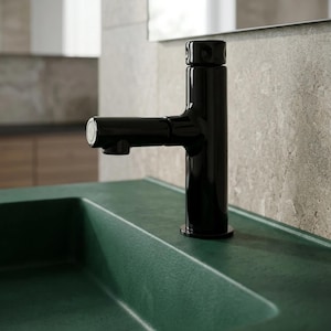 May include: A sleek, glossy black bathroom tap with a cylindrical body and a horizontal spout. The tap features a textured, circular top handle and is mounted directly onto a dark green, matte-finished sink basin with a smooth, curved edge.
