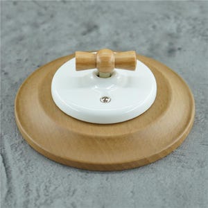Puede incluir: Una placa de pared circular de madera clara con un disco central de cerámica blanca. Un pequeño interruptor de palanca horizontal de madera está montado sobre la base de cerámica blanca, fijada con un tornillo metálico visible en la parte inferior.