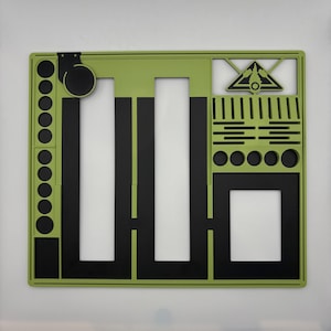 Può includere: Un tappetino rettangolare verde e nero con vari ritagli e forme. Il tappetino presenta una serie di cerchi, rettangoli e linee, creando un design geometrico. La combinazione di colori è verde, nero e bianco.