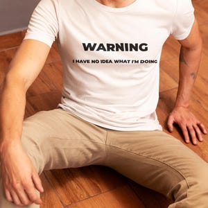 Puede incluir: Camiseta blanca con el texto "WARNING I HAVE NO IDEA WHAT I'M DOING" en negro. La persona lleva pantalones color caqui y está sentada en un suelo de madera.