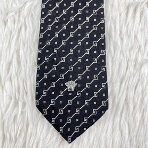 May include: A black tie with a repeating white geometric pattern. The tie features a silver emblem near the bottom and is displayed on a white, textured surface. The tie is a classic accessory.