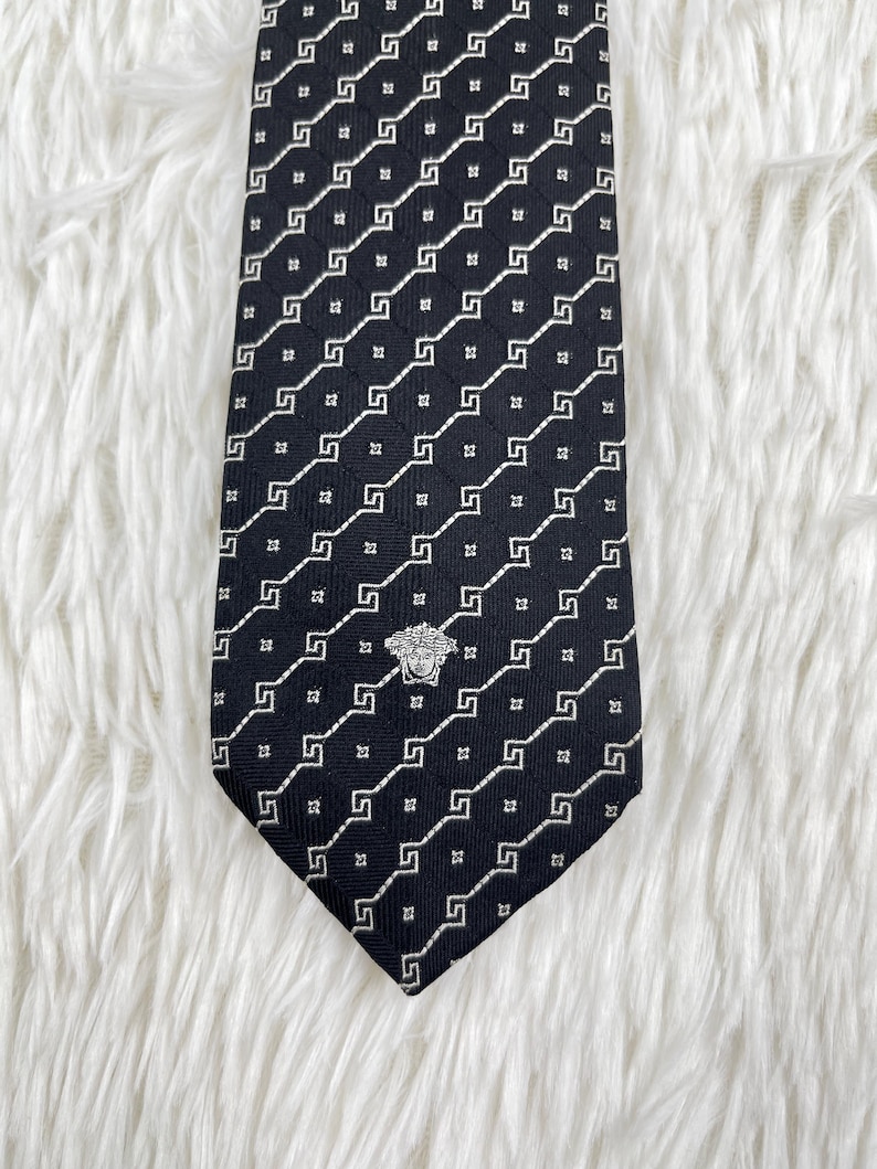 May include: A black tie with a repeating white geometric pattern. The tie features a silver emblem near the bottom and is displayed on a white, textured surface. The tie is a classic accessory.