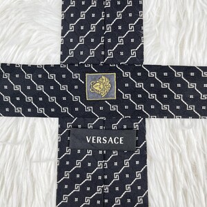 May include: Black silk tie with a repeating white geometric pattern. The tie features a gold and black Versace logo and the brand name "VERSACE" printed on the label.