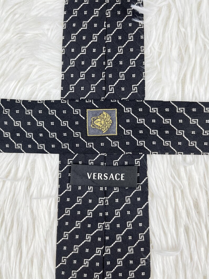 May include: Black silk tie with a repeating white geometric pattern. The tie features a gold and black Versace logo and the brand name "VERSACE" printed on the label.
