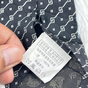 May include: Black Versace tie featuring a white geometric pattern. The fabric has a repeating square and Greek key design. The label displays the Versace logo and care instructions.