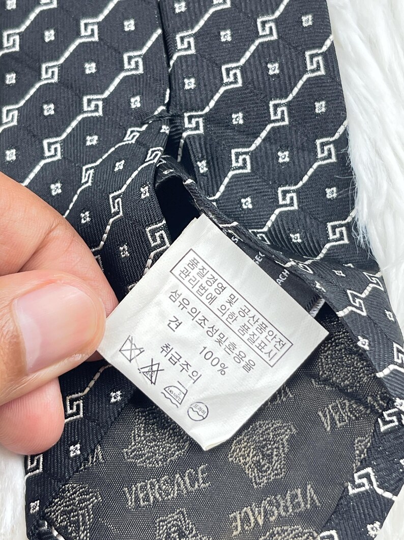 May include: Black Versace tie featuring a white geometric pattern. The fabric has a repeating square and Greek key design. The label displays the Versace logo and care instructions.