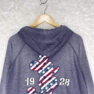 Może przedstawiać: Niebieska bluza z kapturem z grafiką Myszki Miki na plecach. Grafika przedstawia patriotyczny wz&oacute;r z czerwono-białymi paskami i niebieskimi gwiazdami. Widoczny jest r&oacute;wnież tekst "1928" i "SUPER IDOL".