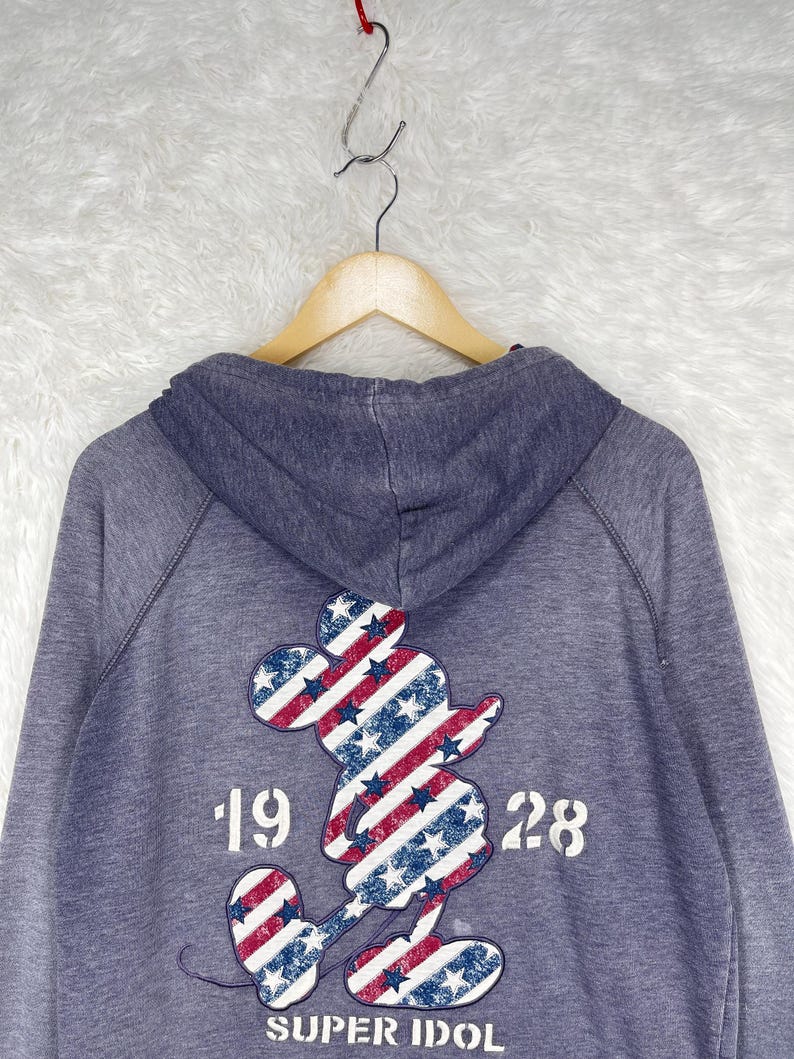 Może przedstawiać: Niebieska bluza z kapturem z grafiką Myszki Miki na plecach. Grafika przedstawia patriotyczny wz&oacute;r z czerwono-białymi paskami i niebieskimi gwiazdami. Widoczny jest r&oacute;wnież tekst "1928" i "SUPER IDOL".