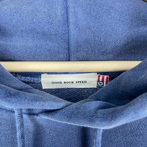 May include: A blue hooded sweatshirt with the text "GOOD ROCK SPEED" on a tag. The sweatshirt is hanging on a wooden hanger. The fabric has a textured appearance.
