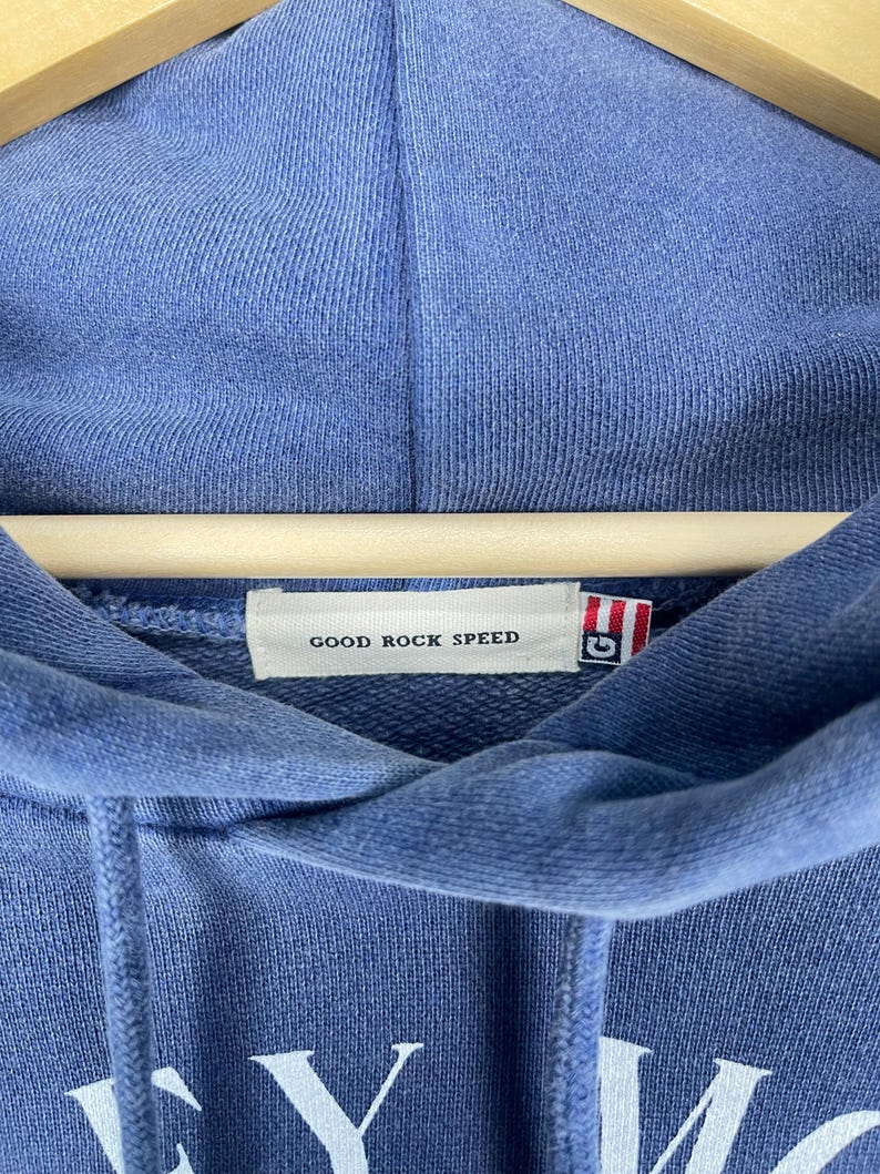 May include: A blue hooded sweatshirt with the text "GOOD ROCK SPEED" on a tag. The sweatshirt is hanging on a wooden hanger. The fabric has a textured appearance.