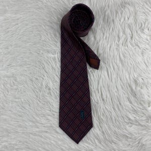 Puede incluir: Una corbata de color burdeos oscuro con un patrón de cuadrícula diagonal repetitivo en líneas finas negras y azul claro. La corbata tiene un extremo puntiagudo y una pequeña etiqueta rectangular cerca de la parte inferior con las letras YSL en azul.