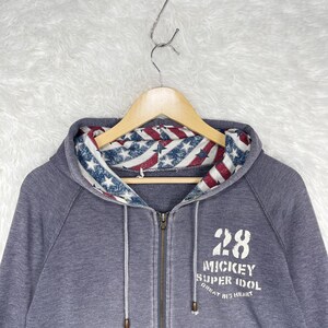 Może przedstawiać: Szara bluza z kapturem zapinana na zamek z kapturem wyłożonym czerwono-biało-niebieskim wzorem w gwiazdy i paski. Bluza ma numer 28 i słowa "Mickey Super Idol Great Big Heart" wydrukowane z przodu.