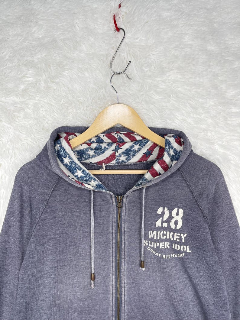 Może przedstawiać: Szara bluza z kapturem zapinana na zamek z kapturem wyłożonym czerwono-biało-niebieskim wzorem w gwiazdy i paski. Bluza ma numer 28 i słowa "Mickey Super Idol Great Big Heart" wydrukowane z przodu.