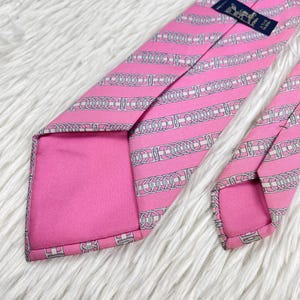 May include: A bright pink necktie featuring a repeating pattern of white and grey interlocking chain links arranged in diagonal stripes. The tie includes a dark blue rectangular label stitched near the top with white text and a horse logo.