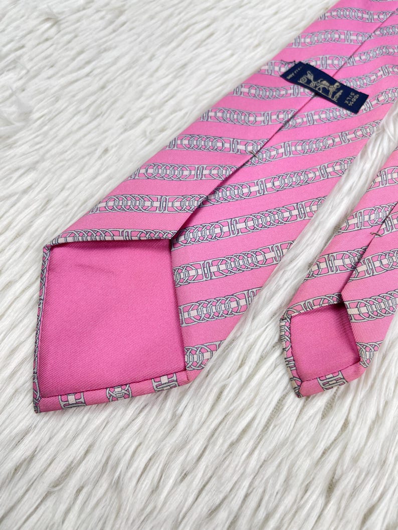 May include: A bright pink necktie featuring a repeating pattern of white and grey interlocking chain links arranged in diagonal stripes. The tie includes a dark blue rectangular label stitched near the top with white text and a horse logo.