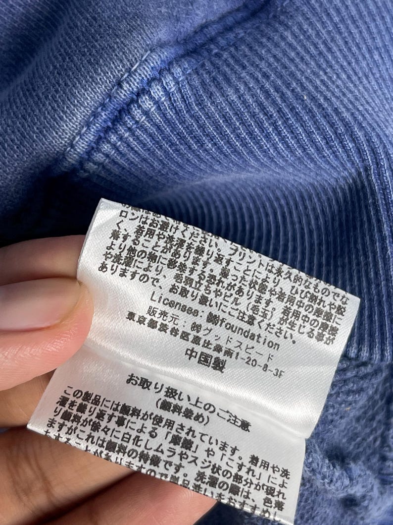 May include: Close-up of a blue fabric with a tag attached. The tag has Japanese text and provides care instructions. The fabric appears to be part of a garment, possibly a sweater or sweatshirt.