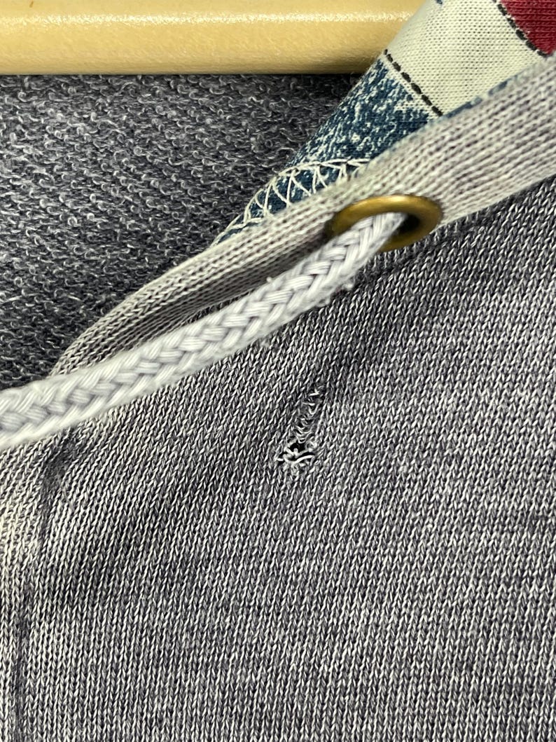 Może przedstawiać: Zbliżenie szarej bluzy z kapturem z białym sznurkiem i wzorzystą podszewką kaptura. Sznurek jest przeciągnięty przez mosiężną przelotkę. W tkaninie znajduje się małe rozdarcie.