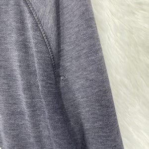 Może przedstawiać: Zbliżenie szarego rękawa bluzy z białym tekstem i małą dziurą. Rękaw ma szew raglanowy i jest wykonany z miękkiej, dzianinowej tkaniny. Tło to biały, puszysty materiał.