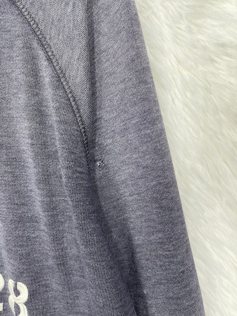 Może przedstawiać: Zbliżenie szarego rękawa bluzy z białym tekstem i małą dziurą. Rękaw ma szew raglanowy i jest wykonany z miękkiej, dzianinowej tkaniny. Tło to biały, puszysty materiał.