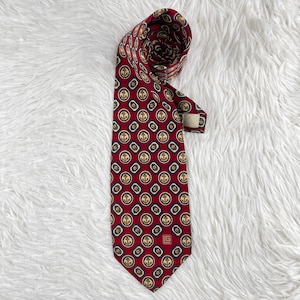 Puede incluir: Una corbata de color rojo intenso con un patrón repetitivo de anclas doradas encerradas en marcos circulares blancos y azul oscuro. La corbata tiene una punta afilada y un pequeño logotipo cuadrado con las letras LG en la parte inferior.