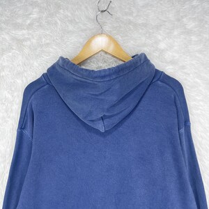 May include: A blue hoodie with a drawstring hood, hanging on a wooden hanger. The hoodie is a solid color, with a slightly faded appearance. The fabric appears to be a medium-weight material.
