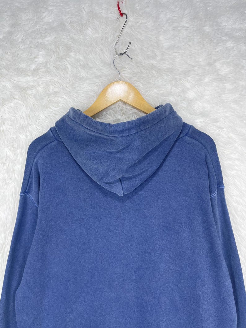May include: A blue hoodie with a drawstring hood, hanging on a wooden hanger. The hoodie is a solid color, with a slightly faded appearance. The fabric appears to be a medium-weight material.